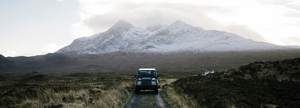4x4 A dark 4x4 off-road vehicle driving down a narrow dirt track toward snow-capped, jagged mountain peaks.
