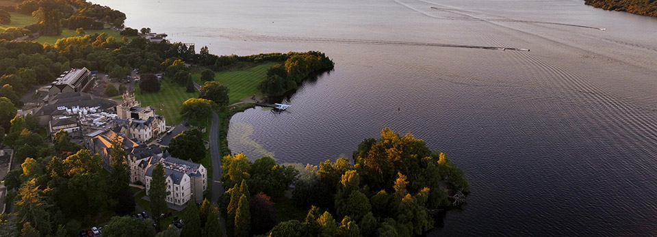 Cameron-House High-angle aerial view of the historic Cameron House hotel nestled along a wooded shoreline next to a large body of water at sunset.