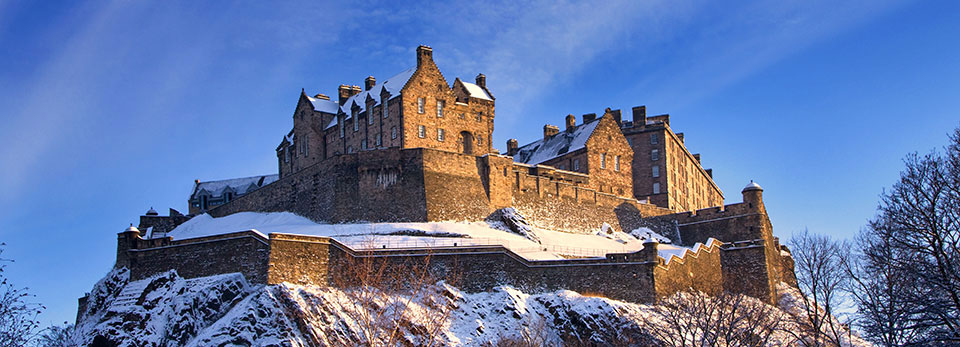 Edinburgh-Castle A wide view of Edinburgh Castle standing atop Castle Rock covered in snow under a clear blue sky.