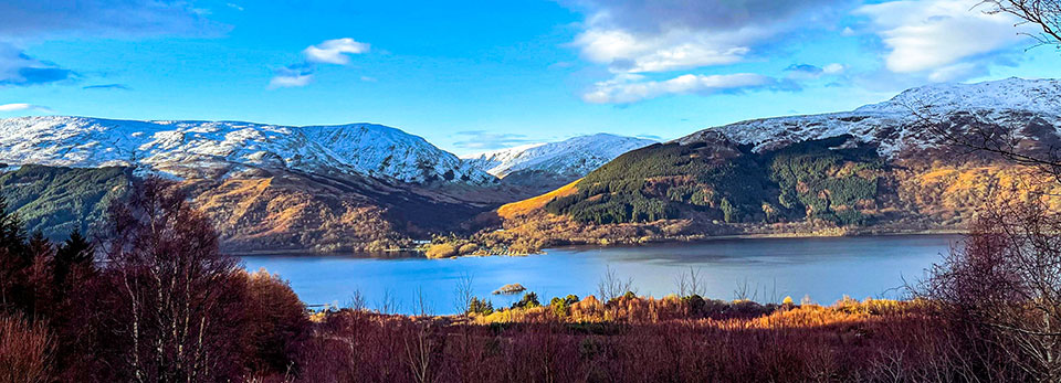 Loch-Lomond A panoramic view across a calm blue loch looking toward snow-capped mountain ridges under a bright blue sky.