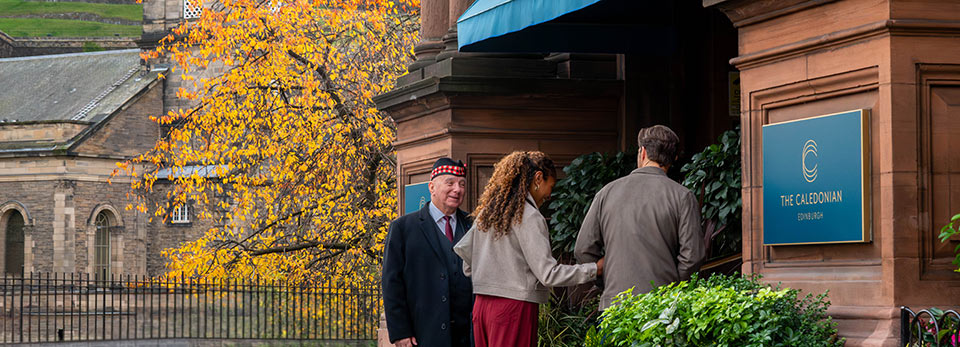 The-Caledonian A couple standing at the entrance of The Caledonian hotel while being greeted by a hotel doorman in traditional Scottish headwear.